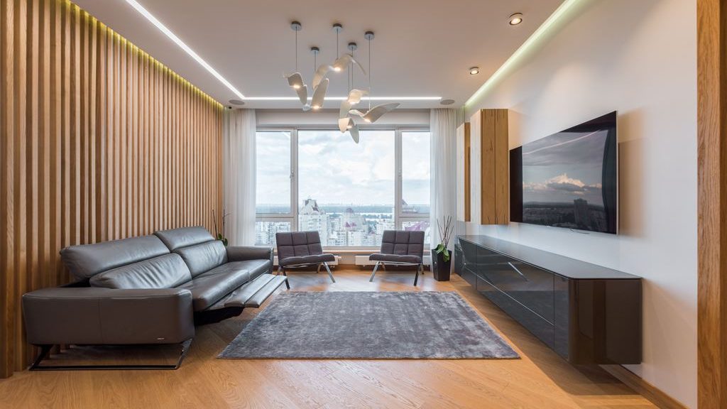 Modern living room with a black leather reclining sofa, gray rug, wall-mounted TV, and floor-to-ceiling city-view windows.