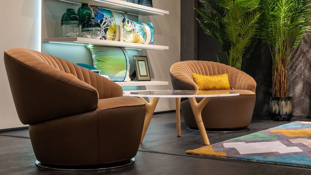 Two brown swivel lounge chairs with quilted backs, arranged around a small glass table in a modern reading corner with illuminated shelves and a tall indoor plant.