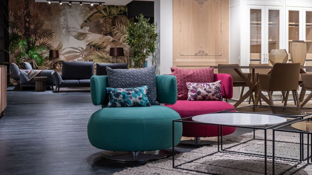 Teal and magenta swivel accent chairs in a contemporary showroom living room, arranged beside black-framed coffee tables and a textured area rug.