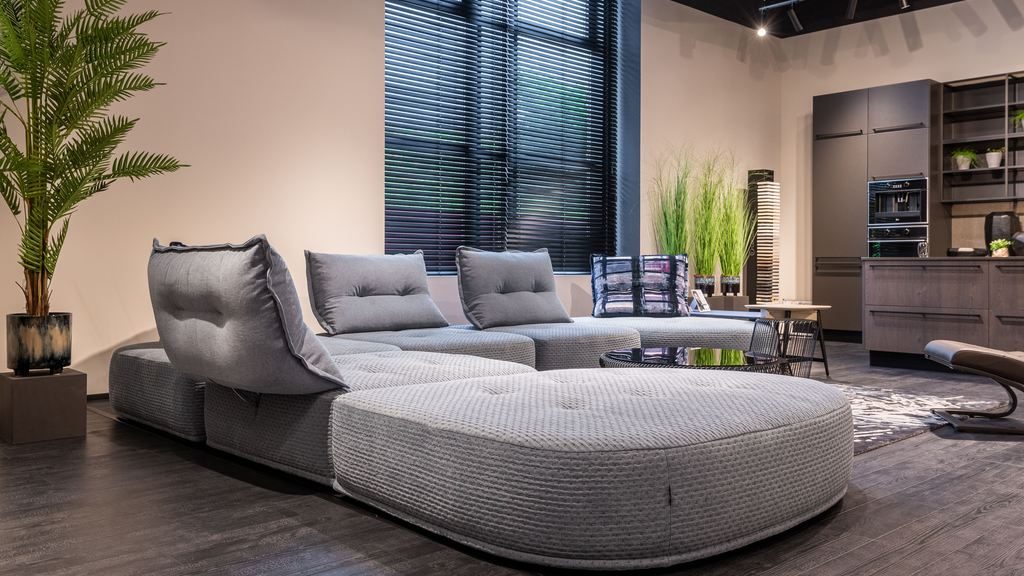 Modern open-plan living room with a low-profile gray modular sectional, oversized ottoman, black blinds, indoor plants, and a dark contemporary kitchen.