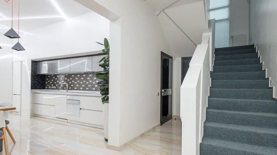 Bright modern interior with carpeted staircase, white railing, open kitchen, pendant lights, and strong diagonal lines guiding the eye upward.
