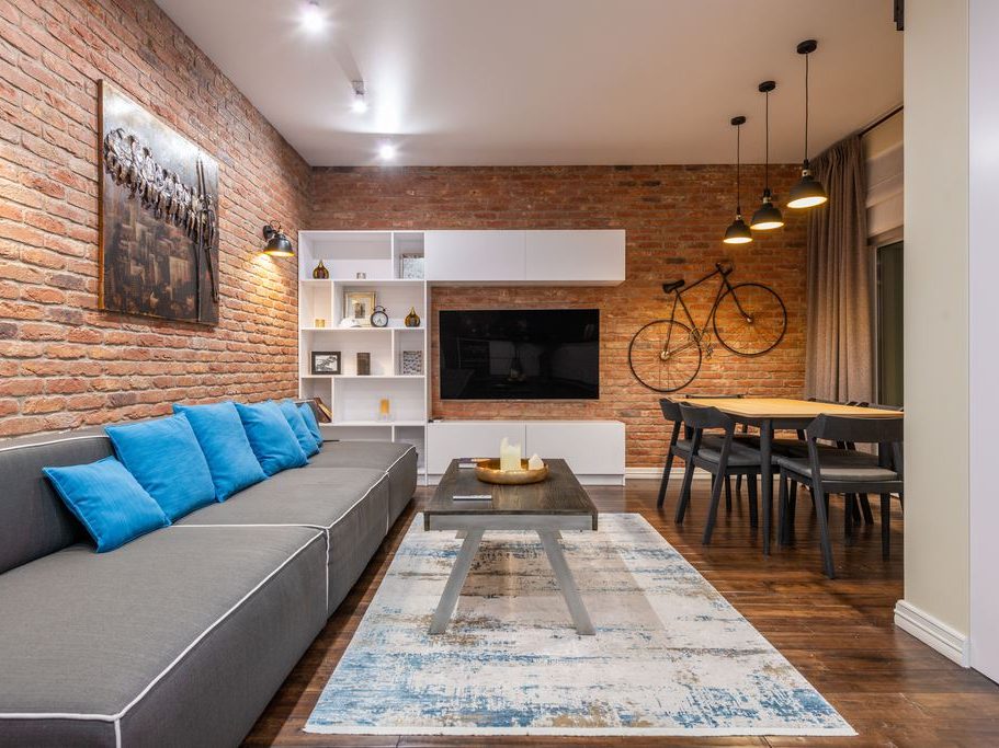 Open-plan living room with a gray sectional sofa, wall-mounted TV, white storage unit, open shelving, and exposed brick walls.