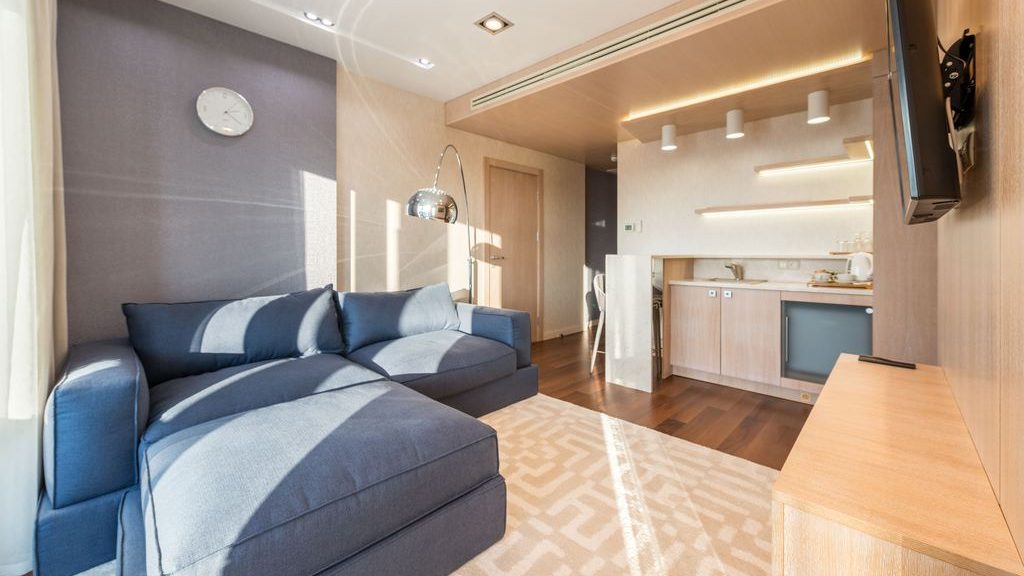 Blue sectional sofa in a compact modern apartment living room with a wall-mounted TV, light wood media console, and open kitchenette.