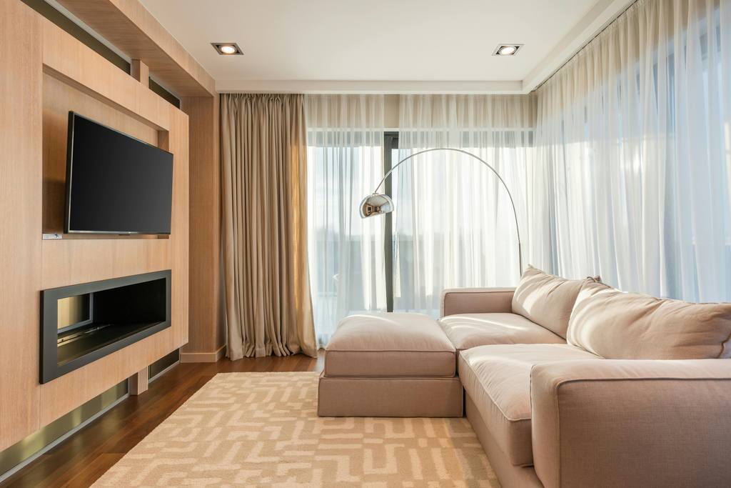 a long and narrow living room with a sofa with an ottoman, a wall-mounted tv and a fire place near a floor lamp