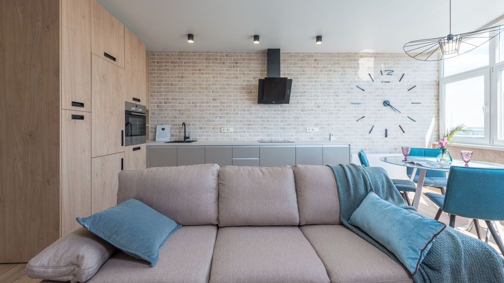 Beige sectional sofa with blue pillows in a bright open-plan apartment, facing a modern kitchen and dining area.