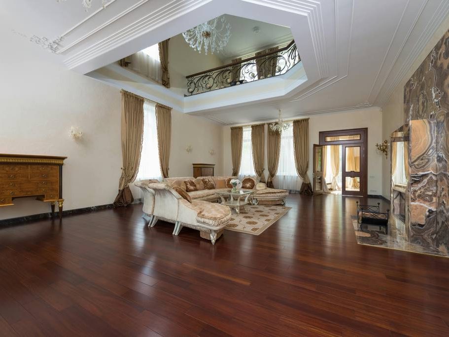 Spacious traditional living room with dark wood floors, cream sectional sofa, ornate fireplace, tall curtains, chandelier, and classic decorative details.