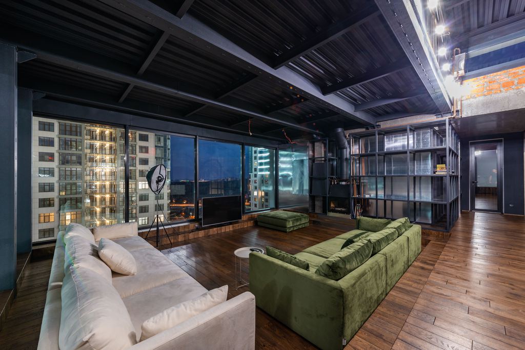 Spacious industrial loft living room with green and white sofas, dark wood floors, floor-to-ceiling windows, and city views at night.