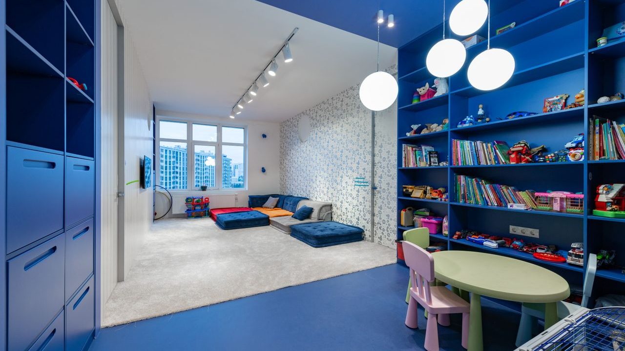 Bright children’s playroom with blue built-in storage, bookshelves, a small activity table, floor cushions, toys, and open play space.