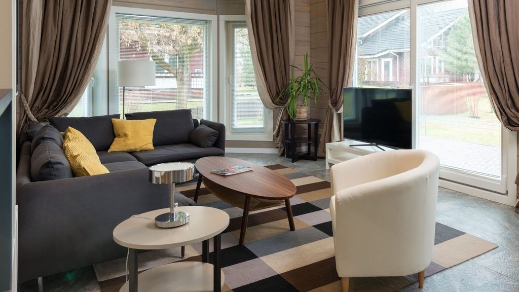 Square living room with a dark sectional sofa, white accent chair, oval coffee table, side tables, and a corner TV by large windows.