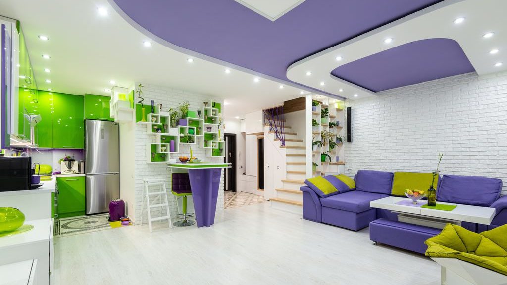 Bright open-plan living room and kitchen with vivid purple seating, lime-green cabinets, geometric ceiling lights, white brick walls, and modern shelving.