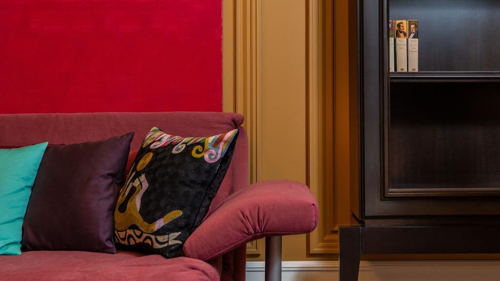 Colorful throw pillows on a red sofa beside a bold abstract painting and dark shelving in a warm, eclectic living room.
