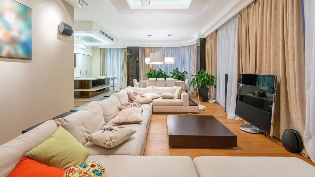 Spacious modern living room with a cream sectional sofa, colorful throw pillows, dark wood coffee table, TV, indoor plants, and floor-to-ceiling curtains.
