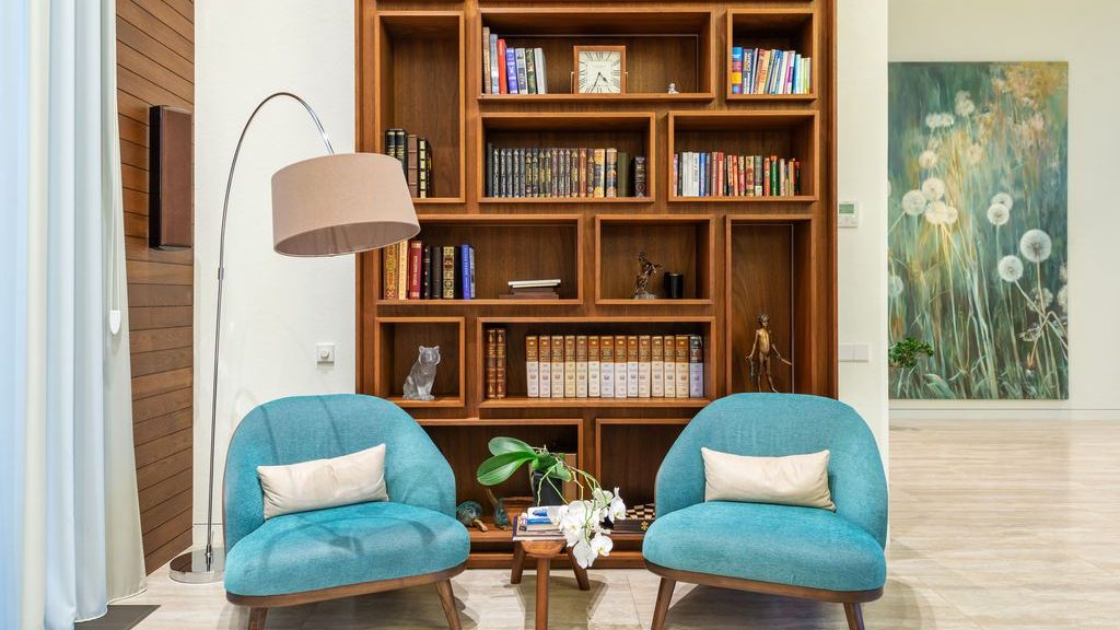 Two teal reading chairs with a small wooden table and floor lamp in front of a built-in wood bookcase.