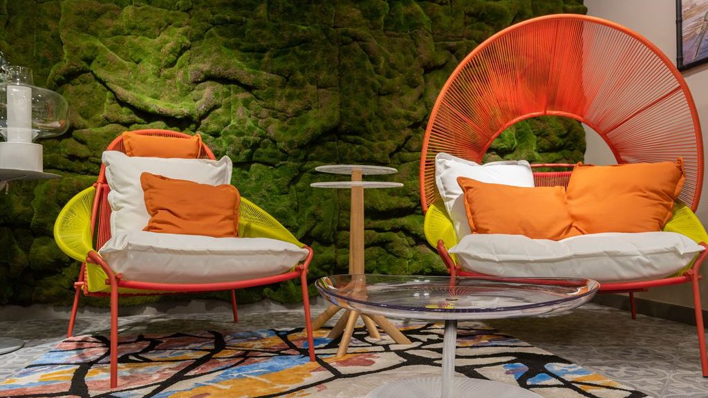 Colorful orange and lime accent chairs with white cushions around a glass coffee table on a patterned rug against a moss-covered feature wall.
