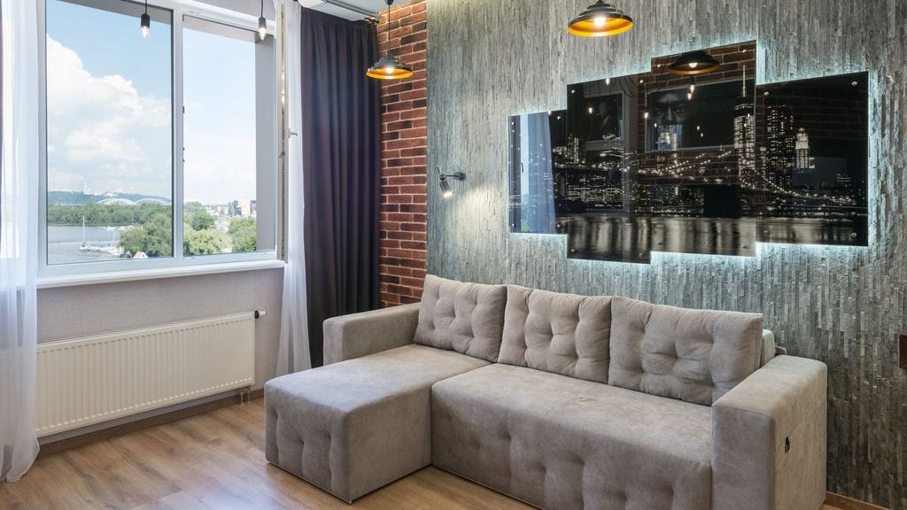 Compact modern living room with a gray sectional sofa, chaise lounge, exposed brick accent wall, and large window.