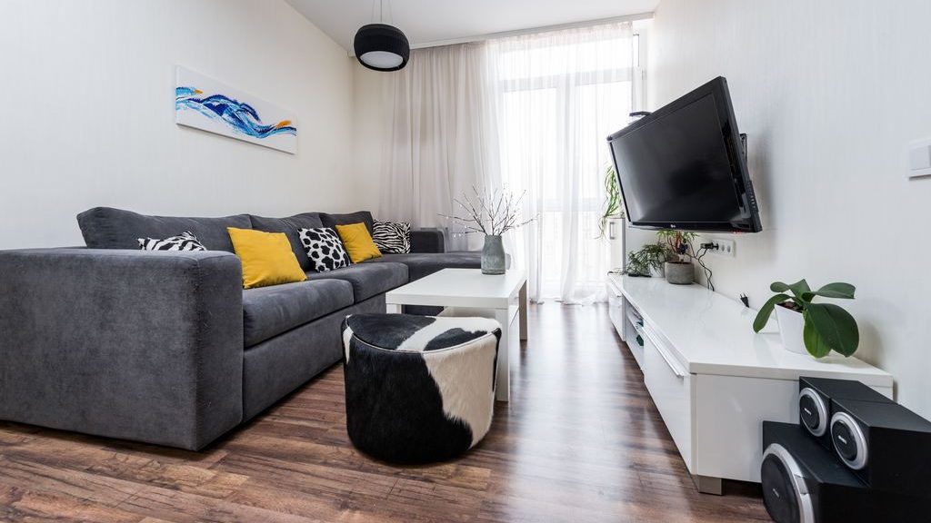Small living room with a gray sofa, wall-mounted TV, white media console, coffee table, and ottoman arranged along a narrow viewing area.