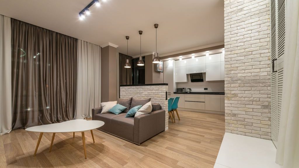 Open-plan living room and kitchen with a gray sofa, oval white coffee table, layered curtains, pendant lights, pale wood flooring, and white brick accents.