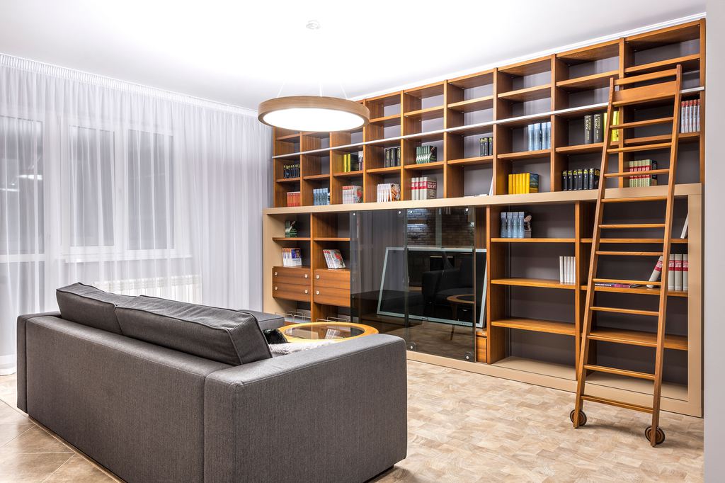 Modern living room with a gray sofa facing a built-in wood bookcase, TV, rolling library ladder, and sheer curtains.