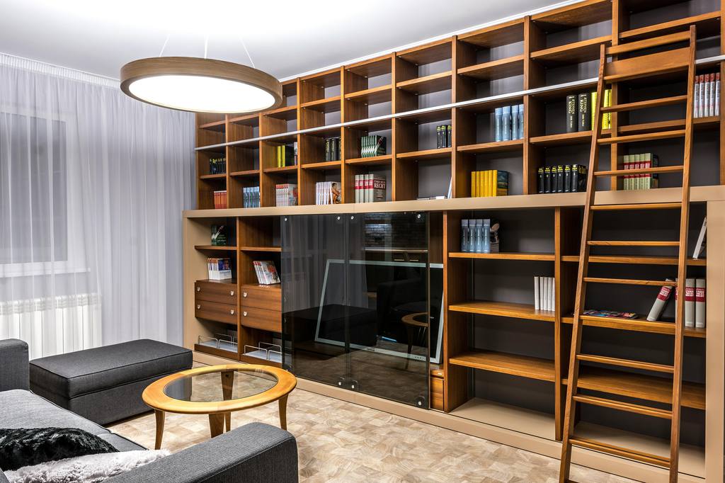 Modern living room with wall-to-wall wooden bookshelves, ladder, glass-front storage cabinets, scattered books, and a low coffee table near a gray sofa.