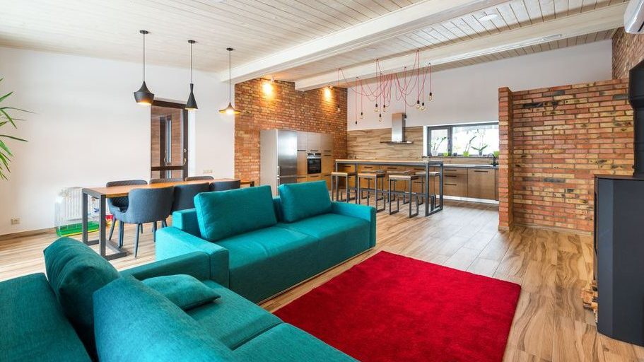Open-plan living room and kitchen with teal sofas, a red area rug, exposed brick walls, pendant lights, and a black wood-burning stove.