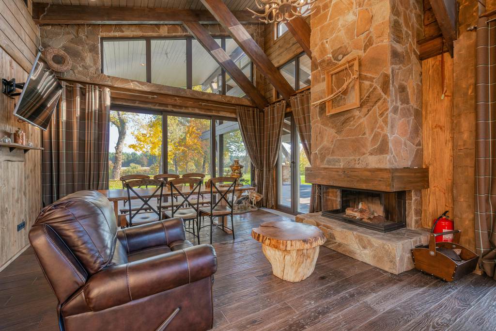 Rustic living room with brown leather recliner, stone fireplace, wood ceiling beams, tree stump coffee table, plaid curtains, and large windows.