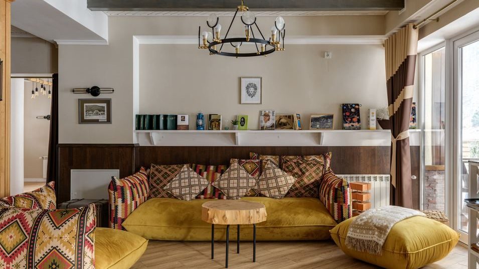 Boho eclectic living room with mustard sofa, patterned pillows, wood coffee table, exposed beams, layered textiles, wall shelf, and warm natural tones.