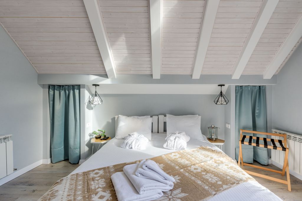 Coastal bedroom with white bedding, light wood ceiling beams, soft blue curtains, neutral throw blanket, and natural wood accents.