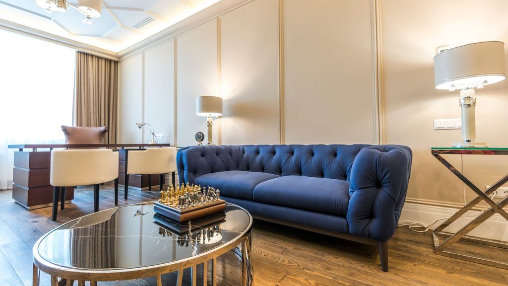 Elegant living room with blue tufted sofa, round glass coffee table, desk area, side lamps, and warm asymmetrical furniture layout.