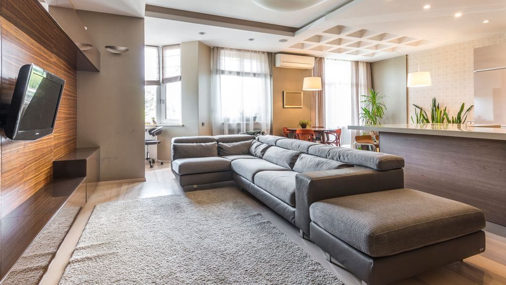 Open-concept living room with a gray reclining sectional, chaise lounge, wall-mounted TV, and adjacent kitchen.