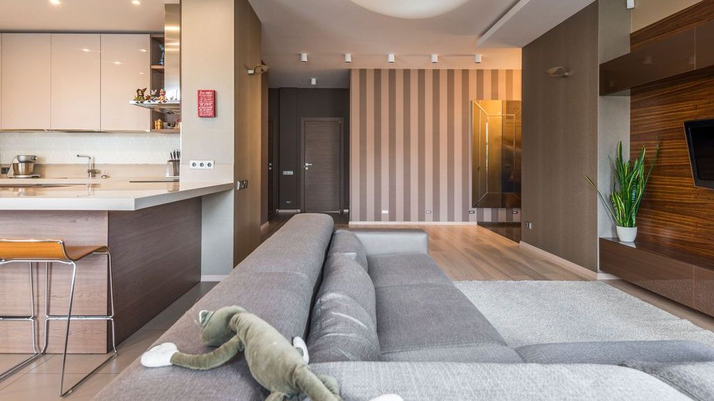 Open-plan rental apartment with gray sectional sofa, kitchen island, striped accent wall, wall-mounted TV, area rug, and warm recessed lighting.