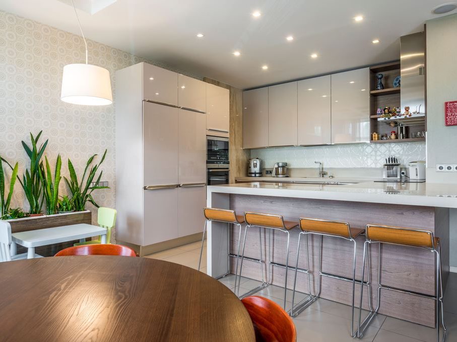 Modern kitchen with island bar seating, built-in coffee station, glossy cabinets, warm wood dining table, pendant light, and indoor plants.