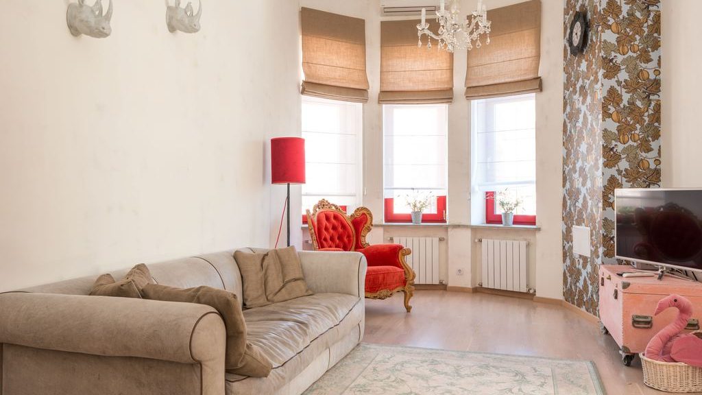 Eclectic living room with a beige sofa, ornate red chair, floral wallpaper, rhinoceros wall decor, pink trunk TV stand, chandelier, and bay windows.