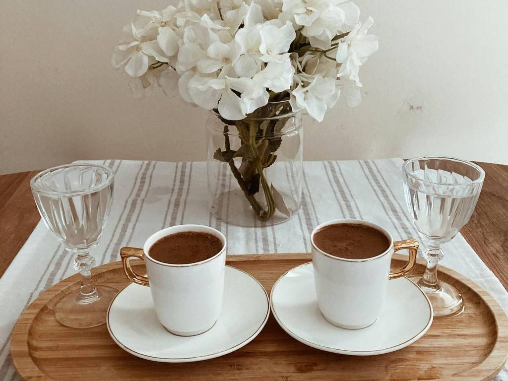 A rustic farmhouse coffee table tray with coffee cups, glassware, and white flowers on a striped neutral tablecloth.