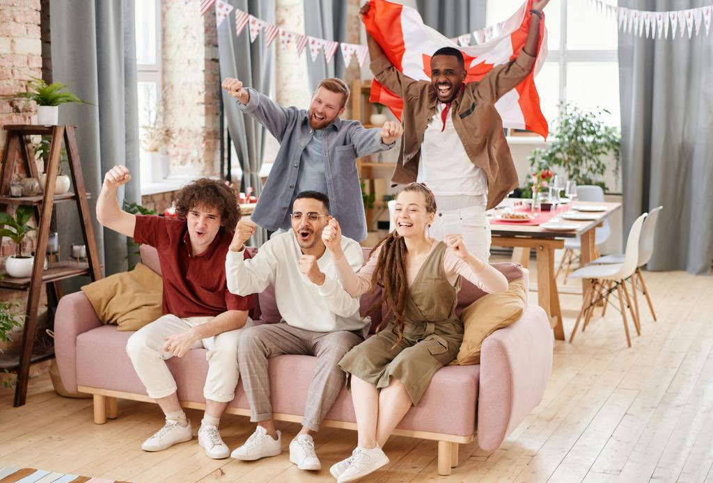 Friends cheering on a pink sofa during a World Cup watch party in a bright living room with dining table and festive decorations.