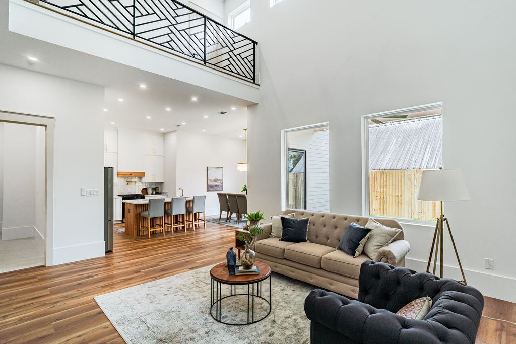 Spacious open-plan living room with tufted sofas, wood floors, kitchen island, dining area, large windows, and high ceilings.