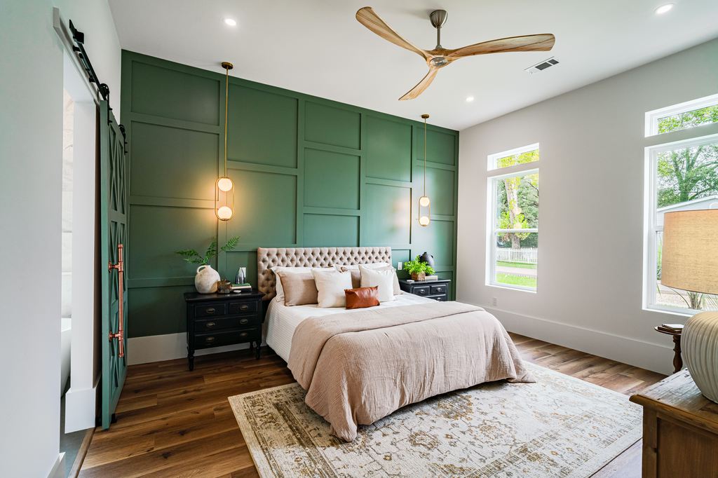 Modern bedroom with green paneled accent wall, tufted upholstered bed, wood flooring, pendant lights, and large windows bringing in natural light.