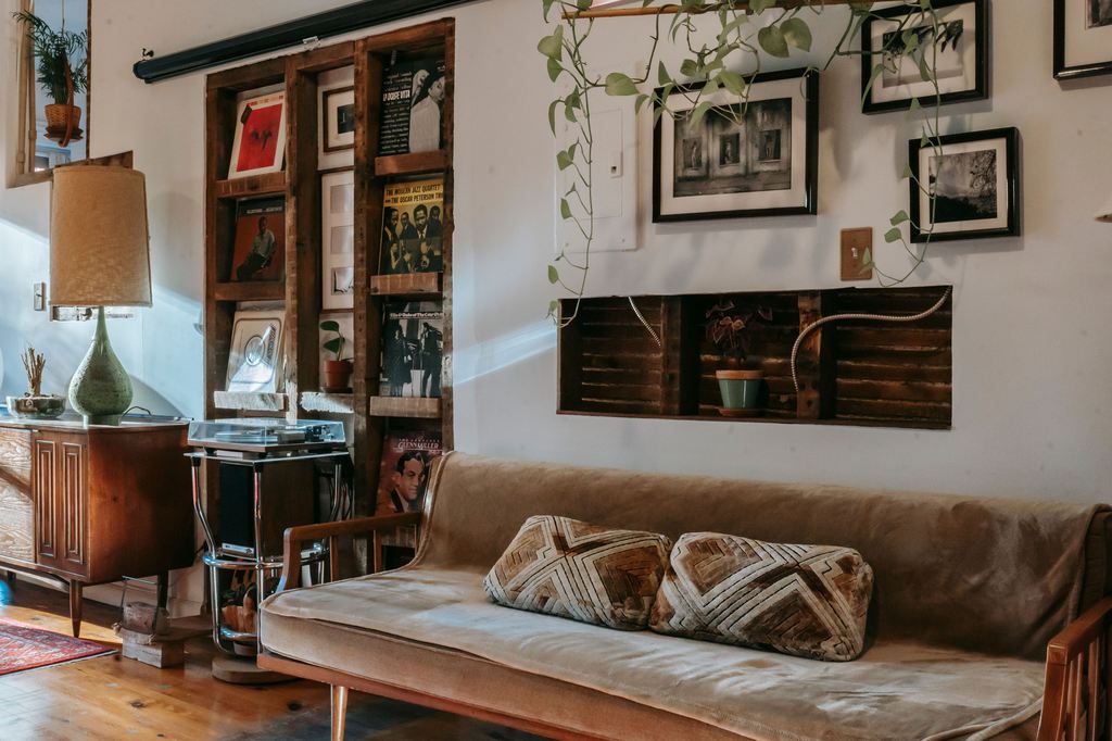 Boho living room with vintage wood furniture, tan sofa, patterned pillows, hanging plants, framed wall art, record shelves, and warm collected decor.