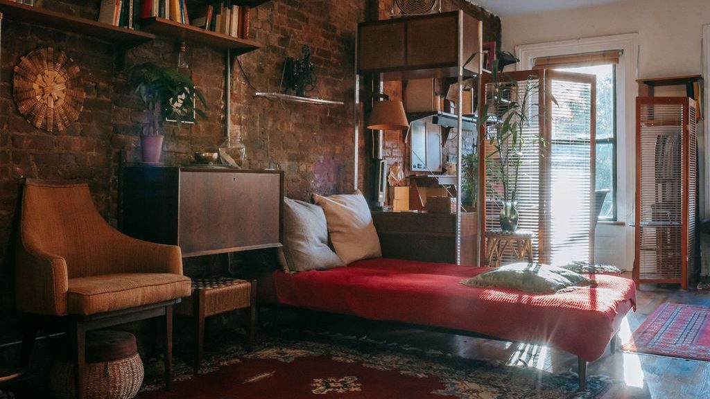 Warm eclectic room with a red daybed, vintage armchair, exposed brick wall, shelving, and a sunlit window.