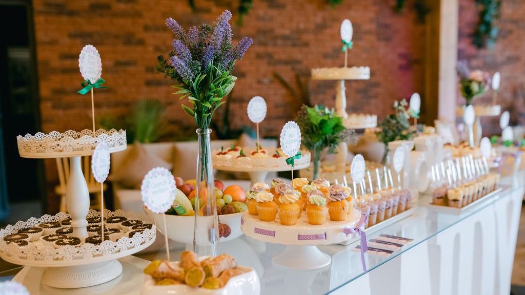 Dessert buffet table with cupcakes, cake stands, fruit, lavender flowers, and labeled treats at a celebration.