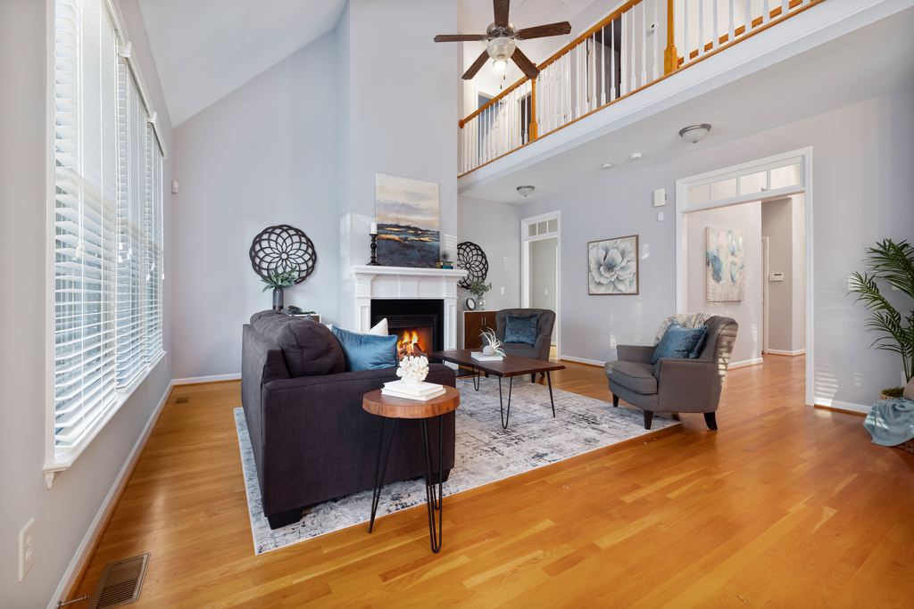 Bright double-height living room with a dark sofa, two gray armchairs, a fireplace, wood floors, and a central rug beneath a loft railing.