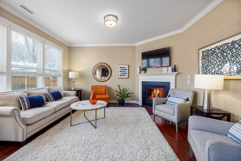 Rental living room with beige sofa, gray accent chairs, large shag rug, fireplace, wall-mounted TV, and layered table lighting.