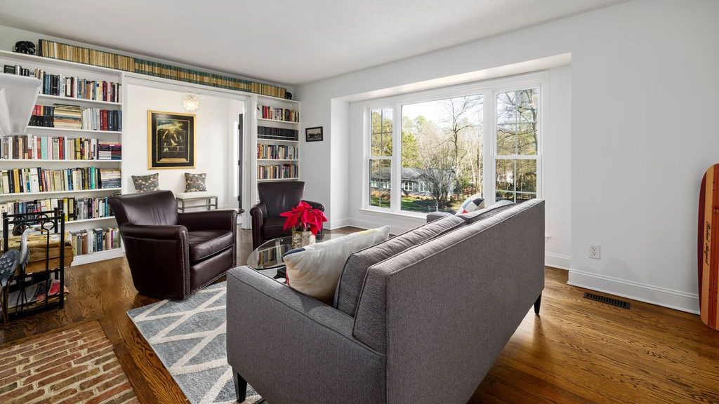 Square living room with a floating gray sofa, two leather armchairs, a round glass coffee table, built-in bookshelves, and a clear window-side walkway.