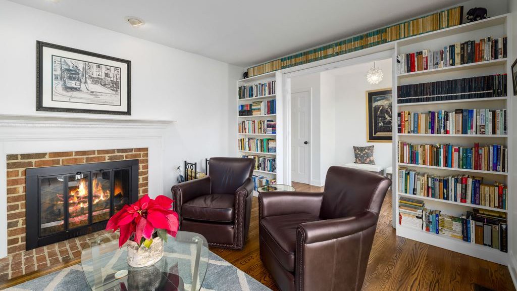 Cozy reading room with two brown leather armchairs, floor-to-ceiling bookshelves, a brick fireplace, glass coffee table, and patterned area rug.