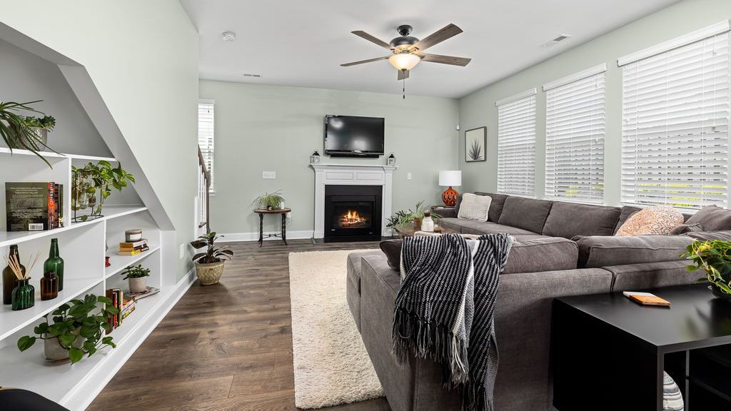 Cozy family room with a large gray sectional, fireplace, wall-mounted TV, built-in shelves, houseplants, and bright windows.