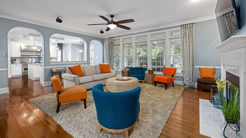 Living room with colorful blue and orange accent chairs, a neutral sofa, round woven coffee table, large area rug, fireplace, and open kitchen view.