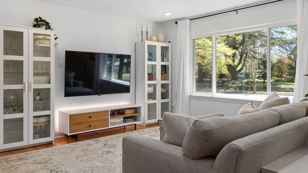 Bright living room with wall-mounted TV above a low TV stand, gray sofa, white storage cabinets, and large window with garden view.