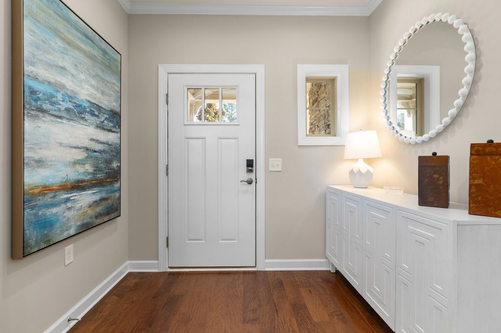 Coastal entryway with white storage cabinet, round shell-style mirror, warm wood floor, table lamp, and abstract ocean artwork.
