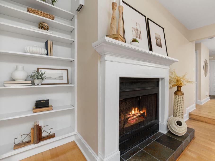 White fireplace with a lit fire, styled mantel art, built-in shelves, light wood flooring, and neutral living room decor.