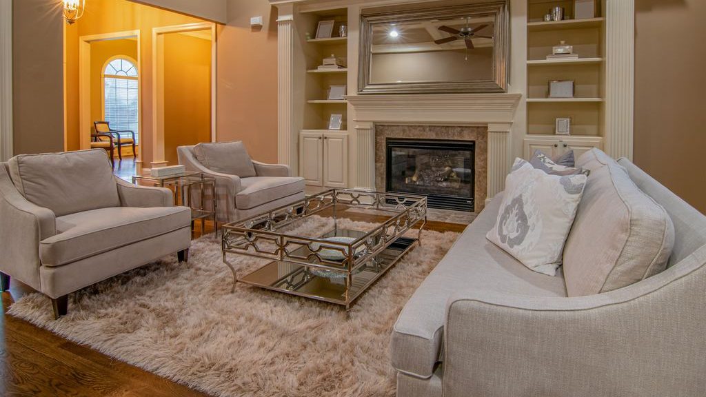 Traditional living room with a beige sofa, two matching armchairs, a mirrored fireplace, built-in shelves, and a shag area rug.