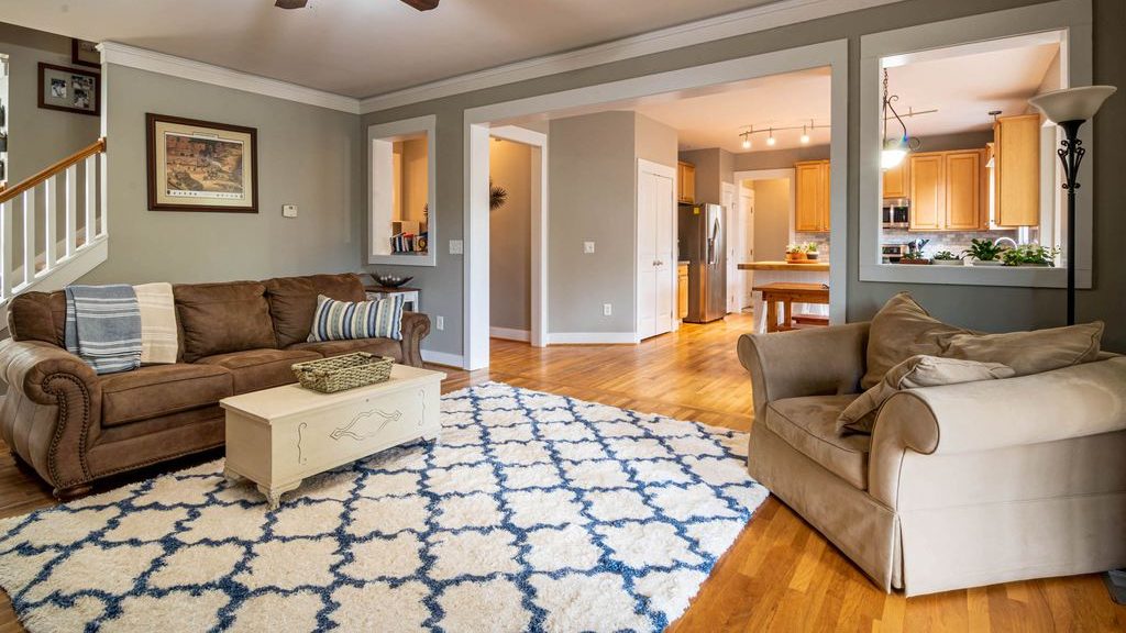 Traditional living room with a brown sofa, beige armchair, patterned blue-and-white area rug, and open views into the kitchen.